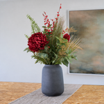 A dark gray ceramic vase filled with an artificial floral arrangement featuring a large deep red flower, red berries, green foliage, and gold grasses, displayed on a table runner in front of a framed abstract painting.