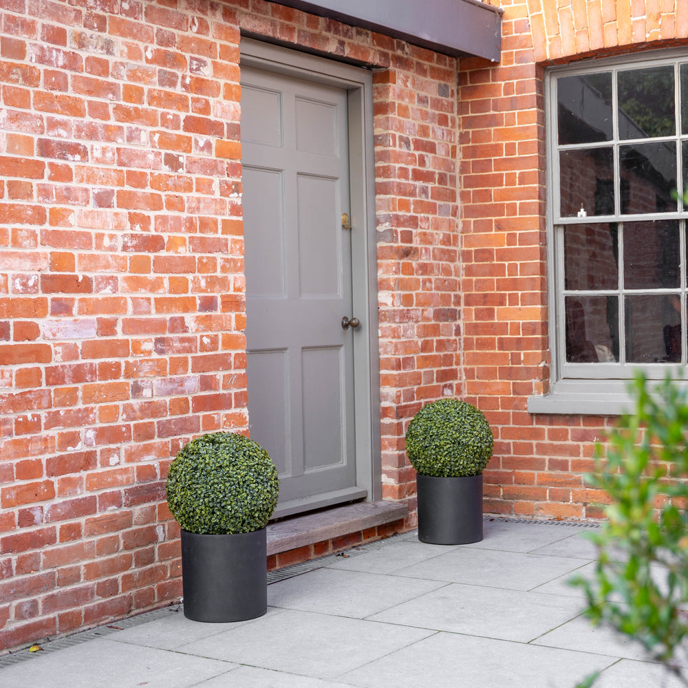 Two spherical green topiary bushes in matte black cylindrical planters placed on either side of a gray door set in a red brick building.