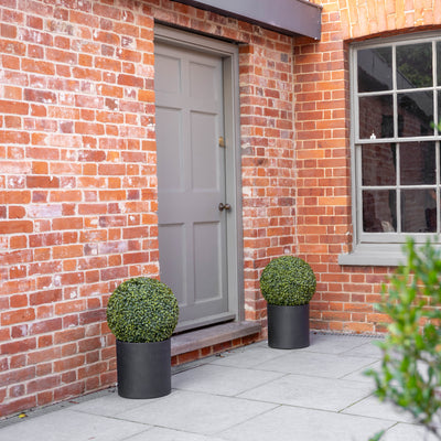 Two spherical green topiary bushes in matte black cylindrical planters placed on either side of a gray door set in a red brick building.