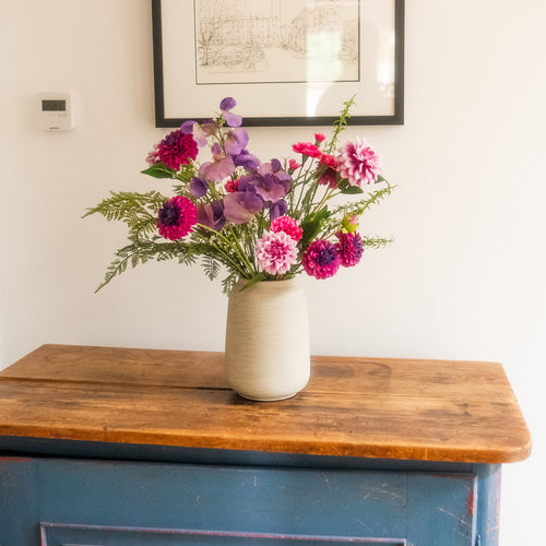 A vase of mixed artificial flowers in pink, purple, and magenta tones with green foliage, displayed on a rustic wooden cabinet against a white wall.