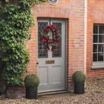 A gray wooden front door with decorative stained-glass panels, adorned with a red holiday wreath. The door is set in a red brick house, flanked by a green ivy-covered wall on the left and a window on the right, with two potted boxwood topiary trees on the gravel ground.