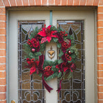 A festive Christmas wreath with red poinsettias, burgundy ribbons, green pine branches, and red berries hangs on a gray front door with decorative stained glass panels, set within a brick wall.