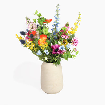 A vibrant and wild artificial flower bouquet in a textured cream-colored ceramic vase, set against a plain white background.