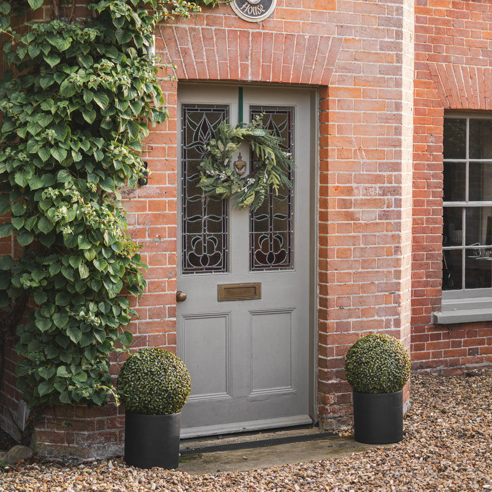A light gray paneled front door with decorative stained glass panels, adorned with a green wreath. The door is set in a red brick building with green ivy on the left, two potted boxwood shrubs on the ground, and a gravel path in front.