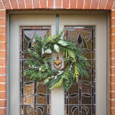 A green holiday wreath with pine branches displayed on a gray front door with decorative stained glass panels, set within a red brick wall.