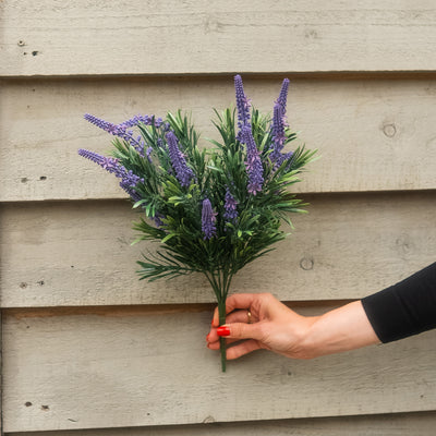 An artificial bouquet of purple flowers with green leaves held by a person's hand against a light gray wooden plank background.