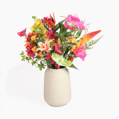 A vibrant artificial flower arrangement featuring tropical blooms with green foliage, presented in a textured off-white ceramic vase against a white background.