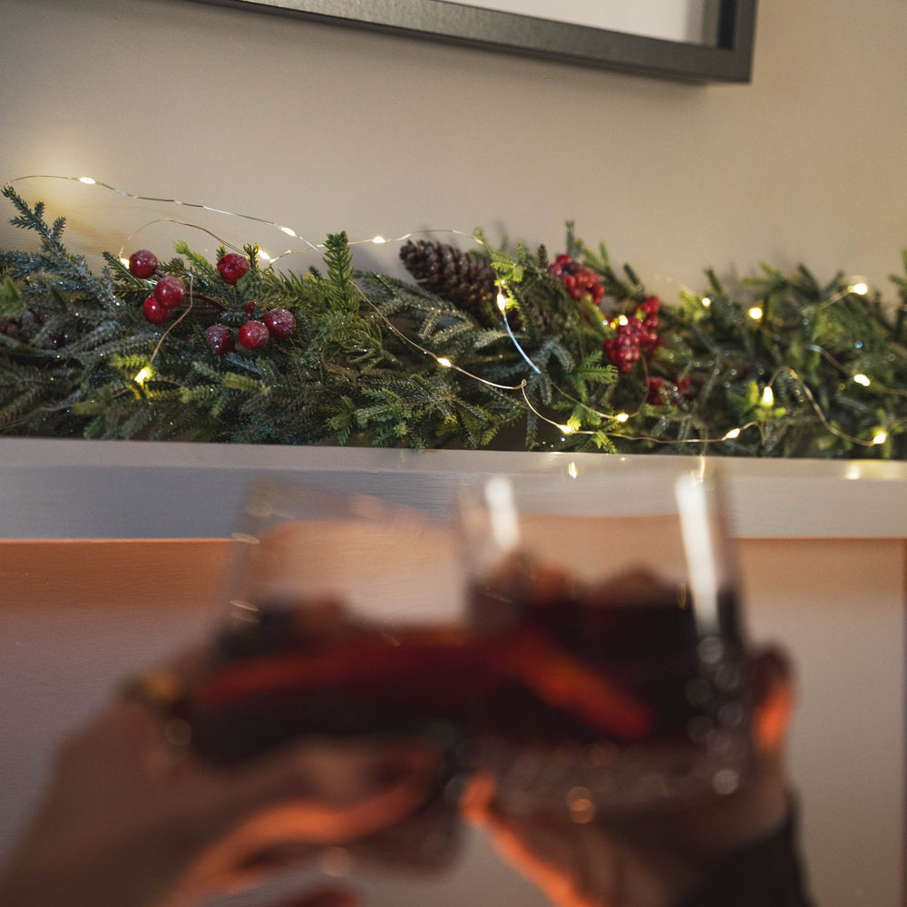 A festive holiday scene with a green garland decorated with pinecones, red berries, and warm string lights across a shelf, while two blurred hands in the foreground hold up glasses of red wine.