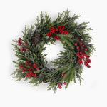 A festive artificial Christmas wreath made of frosted green pine branches, decorated with clusters of red berries and brown pinecones, shown against a plain white background.
