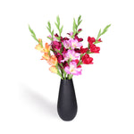 A vibrant artificial flower bouquet featuring pink, orange, and purple gladiolus with green stems, arranged in a matte black teardrop-shaped vase against a solid white background.
