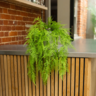 A bright green artificial fern plant hanging over a modern wooden slat counter with a dark metal top, set against a blurred brick wall background.