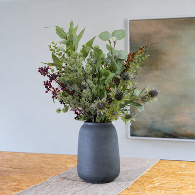 A dark gray ceramic vase filled with an artificial floral arrangement featuring green foliage, purple thistles, and burgundy berries, set on a wooden table with a neutral background and framed wall art.