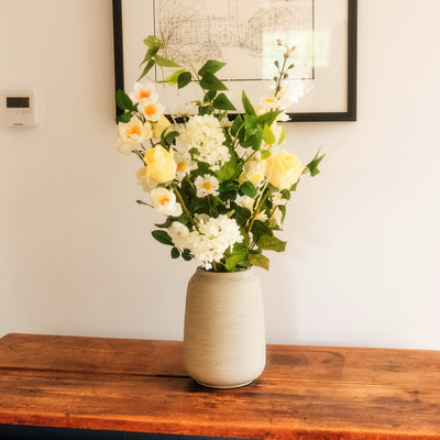 An artificial flower arrangement featuring yellow roses, white daisies, and green foliage in a textured beige ceramic vase, displayed on a rustic wooden table against a white wall.