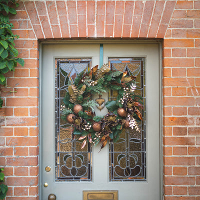 An autumn-themed Christmas wreath with copper ornaments, pinecones, and evergreen branches hanging on a gray front door with stained glass panels, set in a brick doorway.
