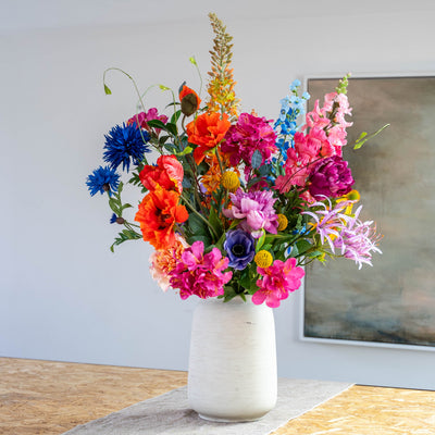 A vibrant, multicolored artificial flower bouquet arranged in a textured white ceramic vase, set on a wooden table against a neutral wall with a framed painting.