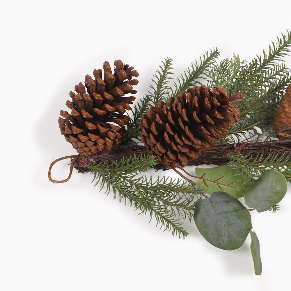 A close up of the loop at the end of a decorative garland featuring pinecones, green pine needles, and round green eucalyptus leaves on a white background.