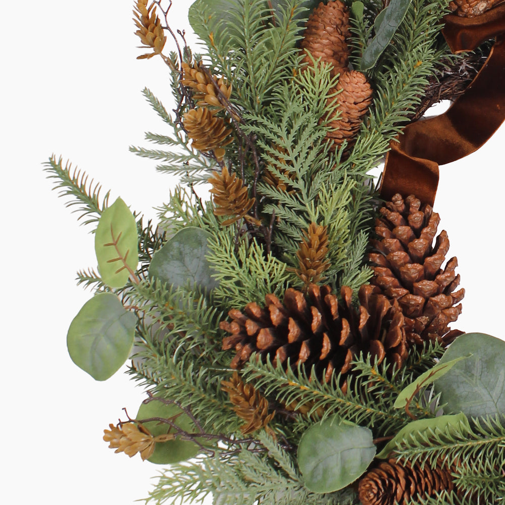 A close-up of a rustic pine wreath featuring green fir and pine branches, natural brown pinecones, and a wide brown velvet ribbon, set against a plain white background.