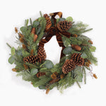 A festive pine wreath decorated with green pine needles, brown pinecones, eucalyptus leaves, and a large brown velvet bow, photographed against a clean white background.
