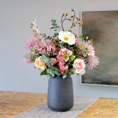 A full floral arrangement in a matte dark gray vase, featuring pink, peach, and white flowers with a touch of green foliage, displayed on a table runner against a neutral wall.