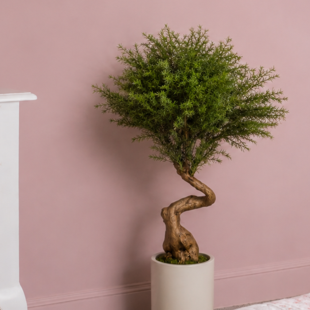 An artificial bonsai-style green tree with a twisted brown trunk in a white cylindrical pot, set against a dusty pink wall.
