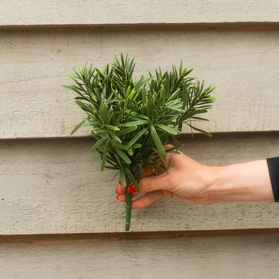 A hand holding a small bunch of green artificial leaves against a beige exterior wall background.