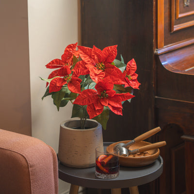 A red artificial poinsettia plant in a grey textured pot, styled on a dark gray side table with a glass and a wooden bowl of walnuts next to a piano.