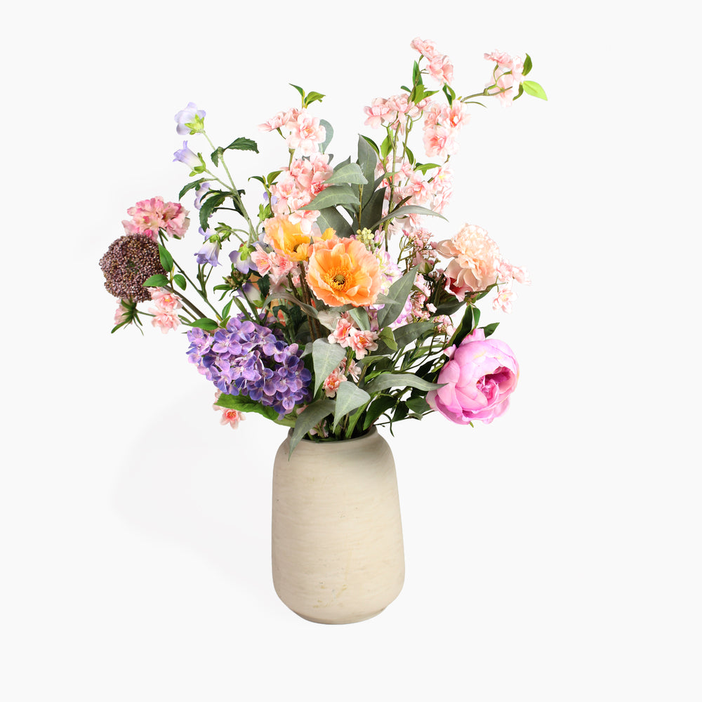 A mixed artificial flower bouquet with tall trailing blossom and a mixture of large and small flower soft coloured flowers in a textured beige ceramic vase, set against a white background.