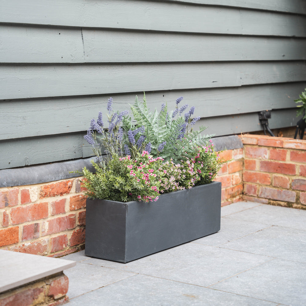 Tall rectangular fiberclay planter with purple and pink flowers and foliage on a patio next to a house exterior.