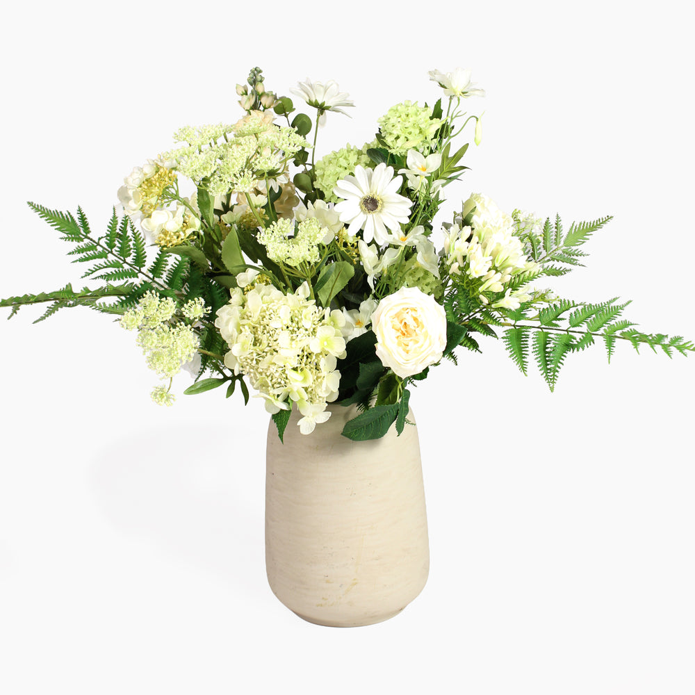 An artificial floral arrangement featuring white and cream flowers, green foliage, and ferns, presented in a textured beige ceramic vase against a clean white background.