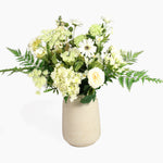 An artificial floral arrangement featuring white and cream flowers, green foliage, and ferns, presented in a textured beige ceramic vase against a clean white background.