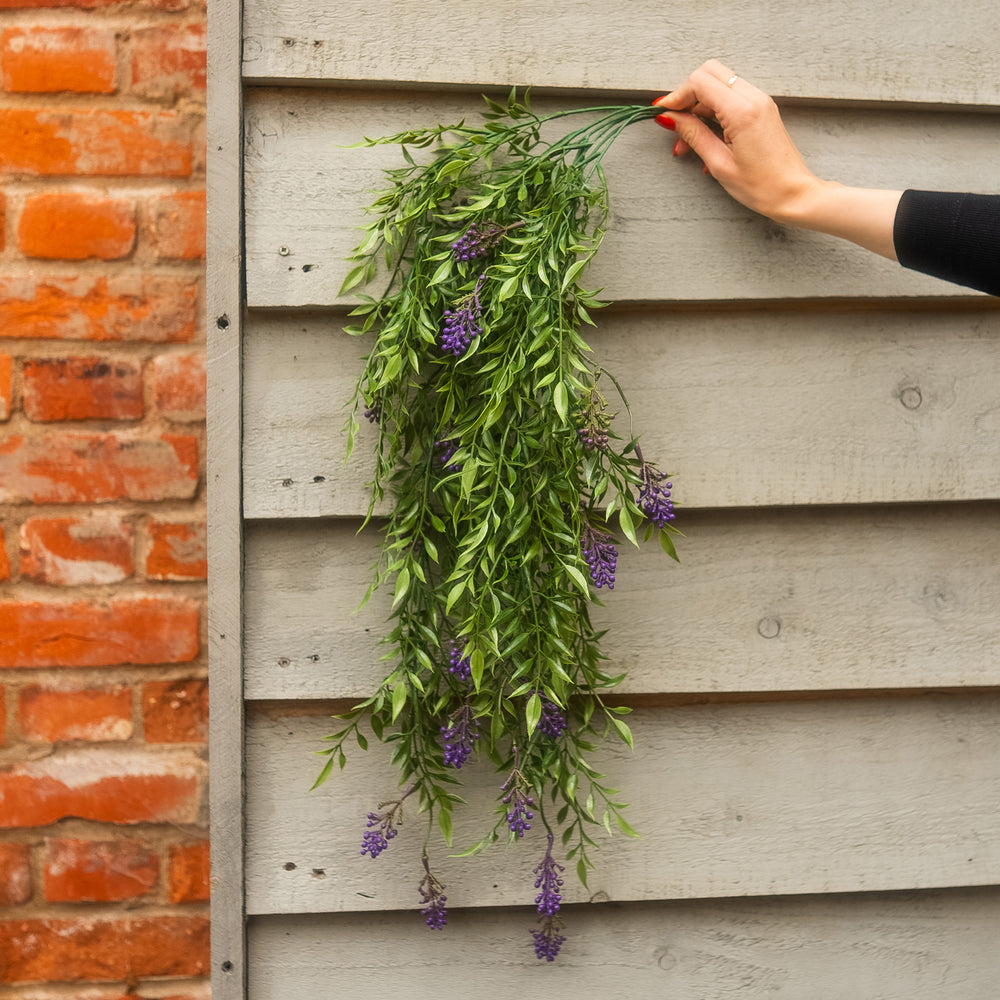 A person's hand with red nail polish holds a bunch of artificial green leaves and purple wisteria flowers against a gray wooden siding wall next to a red brick wall.
