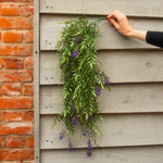 A person's hand with red nail polish holds a bunch of artificial green leaves and purple wisteria flowers against a gray wooden siding wall next to a red brick wall.