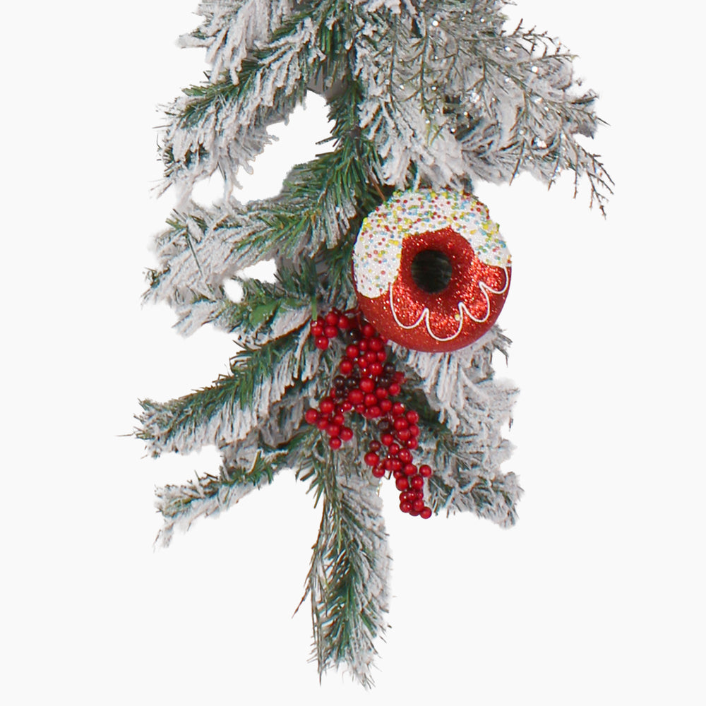 A festive Christmas garland branch decoration featuring a frosted pine with a red and white donut ornament, white icing details, and clusters of red berries, isolated on a white background.