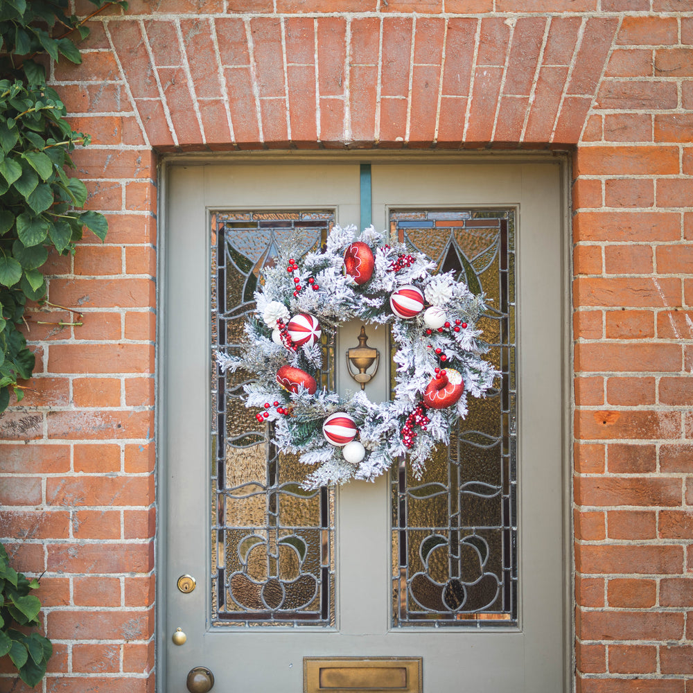 A festive Christmas wreath with white faux snow, red ornaments, and berries hangs on a gray front door with stained glass panels, set against a red brick wall.