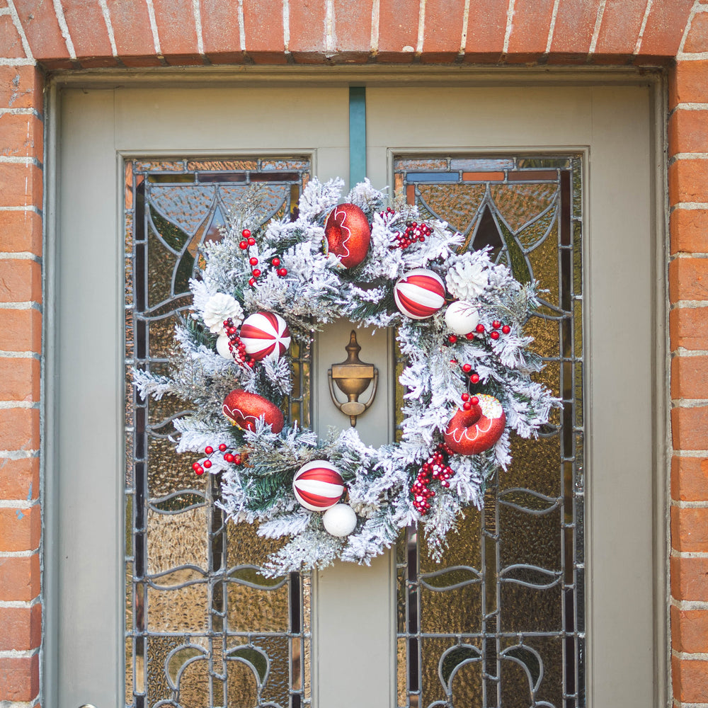 A festive Christmas wreath with white faux snow, red ornaments, and berries hangs on a gray front door with stained glass panels, set against a red brick wall.