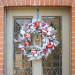 A festive Christmas wreath with white faux snow, red ornaments, and berries hangs on a gray front door with stained glass panels, set against a red brick wall.
