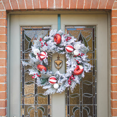 A festive Christmas wreath with white faux snow, red ornaments, and berries hangs on a gray front door with stained glass panels, set against a red brick wall.