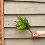 A hand with red nail polish holds a green artificial fern bouquet against a light gray wooden siding background.