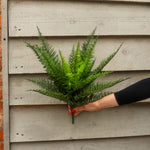 A person's hand with red nail polish holds a green artificial fern bouquet against a light gray wooden siding background.