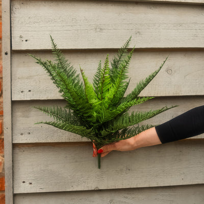 A person's hand with red nail polish holds a green artificial fern bouquet against a light gray wooden siding background.