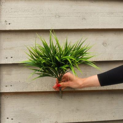 A hand with red nail polish holds a small artificial green plant with long, slender leaves against a light gray wooden plank background.