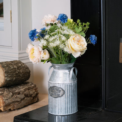 Decorative floral arrangement in a metal vase on a fireplace hearth with logs.