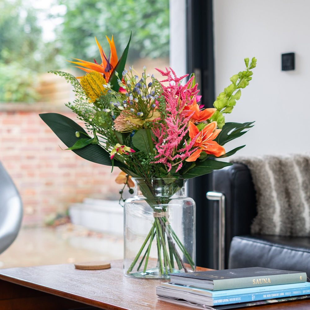 Floral arrangement in a glass vase on a wooden table with a couch and window in the background