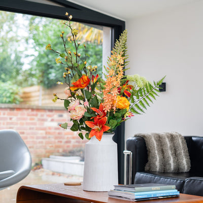 Floral arrangement on a wooden coffee table in a living room with a couch and window in the background.