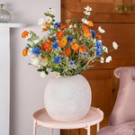 Colorful bouquet of flowers in a white vase on a small table next to a pink chair.