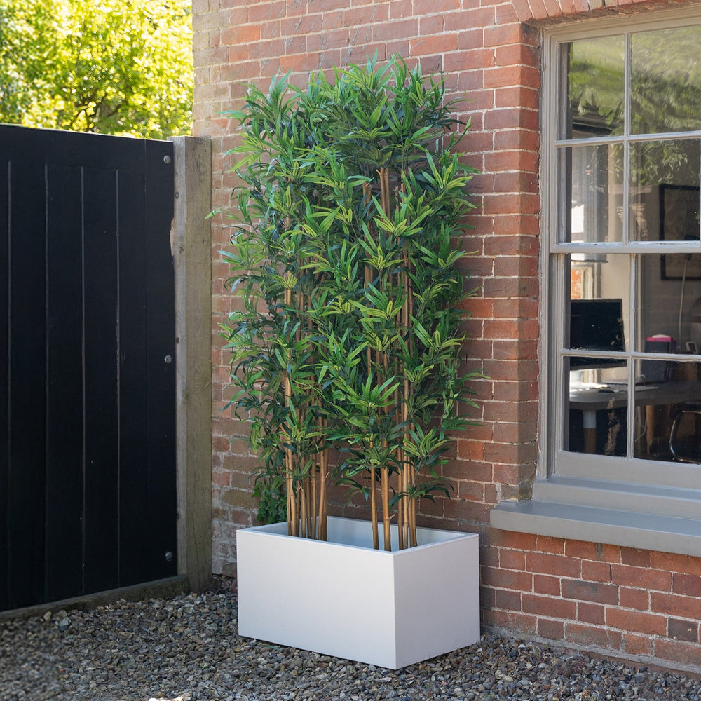 Artificial bamboo plant in a white planter against a brick wall.