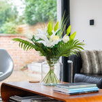 Bouquet of white flowers and green leaves in a clear vase on a wooden table with books, in a room with a window and sofa.