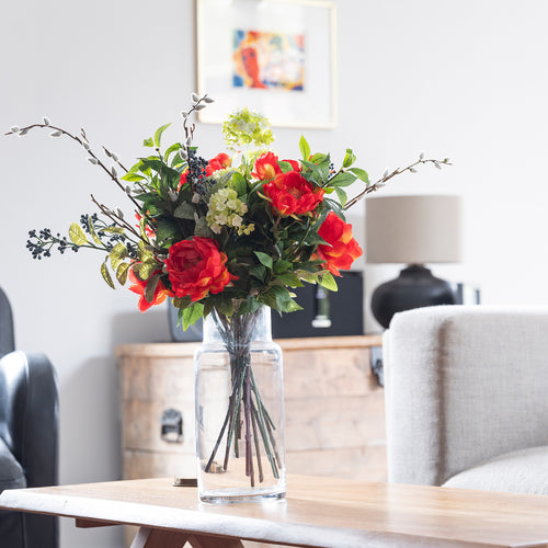 Bouquet of red and green flowers in a clear vase on a wooden table in a living room.