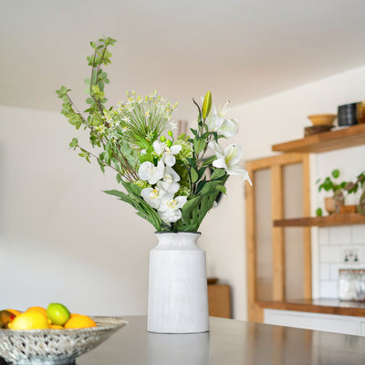 White vase with greenery and white flowers on a table in a room with wooden shelves.