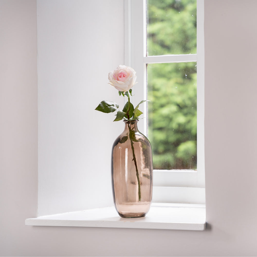 Vase with a single pink rose on a windowsill with a blurred green outdoor background